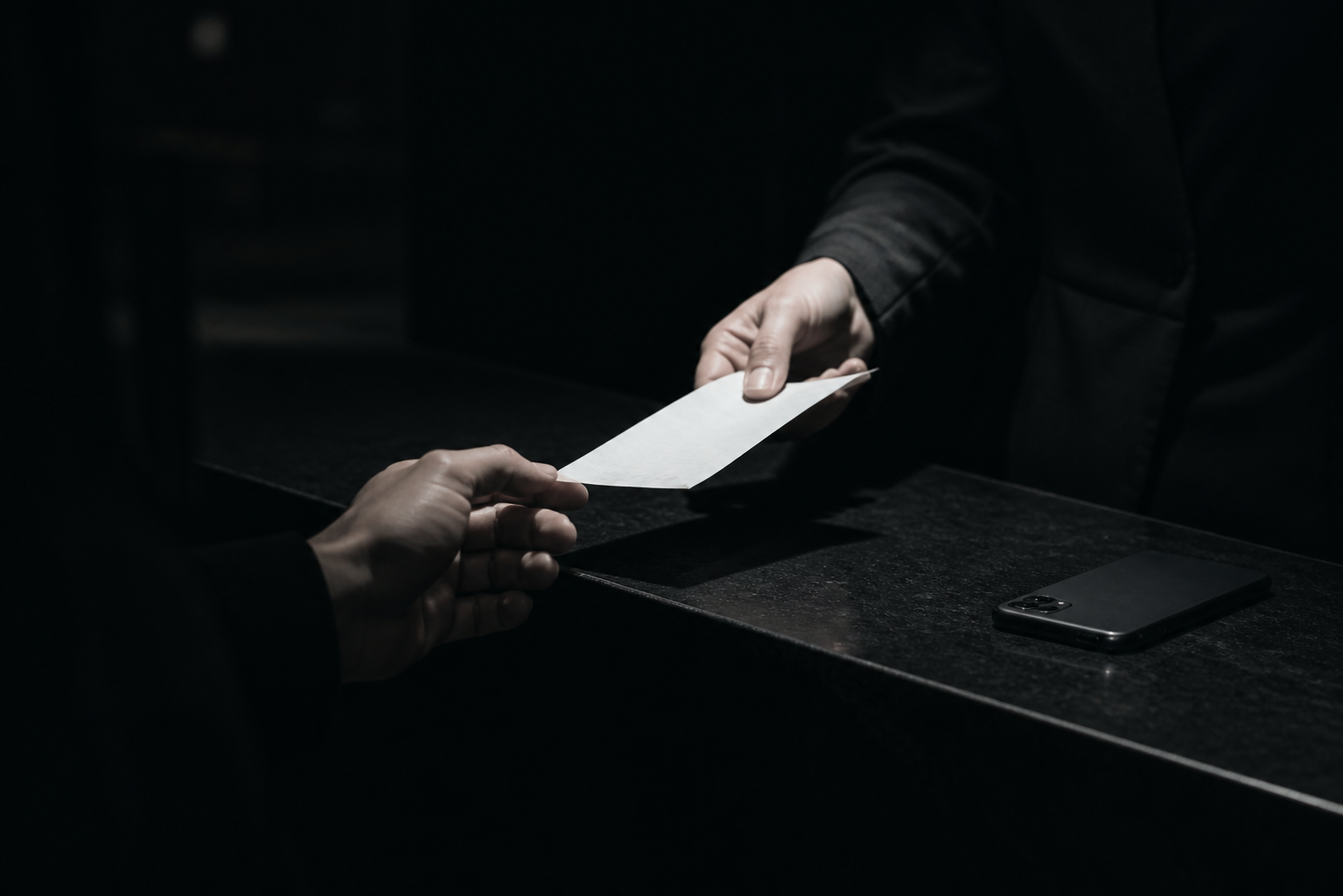 A hand exchanging a paper receipt under a harsh white light at a checkout counter, cold and monochrome, with a phone lying face-down and unlit nearby